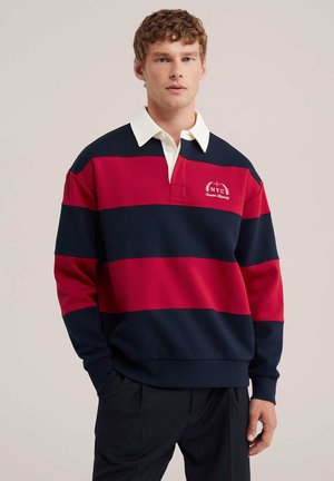 Jeune homme aux cheveux châtain clair bouclés portant un maillot de rugby rayé bleu marine et rouge avec col blanc et logo NYC brodé.