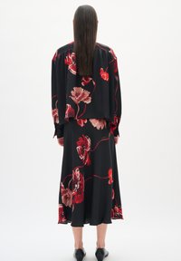 Donna in piedi di spalle, con una blusa nera e una lunga gonna con grandi motivi floreali rossi e rosa, e scarpe nere.