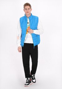 Gilet blu gonfiabile con colletto alto, chiusura con zip e dettagli verdi. Indossato sopra una felpa bianca con grafica e pantaloni neri, abbinato a sneaker nere.
