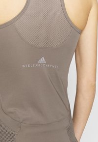 Brun atletisk linne med nätpanel, prytt med vita Adidas- och Stella McCartney-logotyper. Slät textur med en figurnära design.