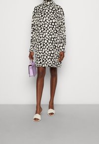 Zwarte jurk met witte bloemenprint, lange mouwen en hoge hals. Gecombineerd met een lavendelkleurige handtas en lichtgekleurde slide sandalen.