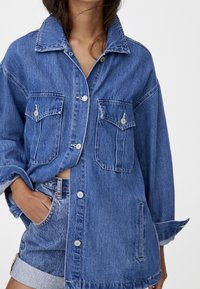 Mujer vestida con una chaqueta de mezclilla azul con bolsillos abotonados y pantalones cortos de mezclilla de cintura alta a juego, con la mano en el bolsillo, de pie contra un fondo blanco.