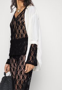 Un haut en dentelle noire avec des manches longues et un décolleté en V, superposé sous une chemise blanche en soie ouverte. Un pantalon en dentelle noire et un sac à main gris texturé complètent le look.