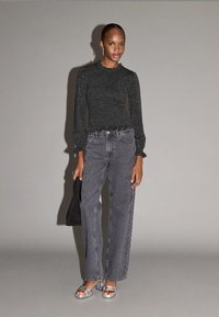 Top nero luccicante a maniche lunghe con orlo arricciato, abbinato a jeans grigi a vita alta e gamba dritta e sandali argentati con tacco. Borsa nera in mano.