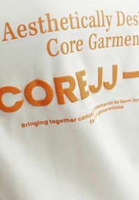 White fabric with orange text. Features the phrases "Aesthetically Designed Core Garments" and "COREJJ—" in bold fonts.