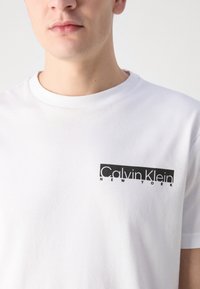 Balta medvilnės marškinėliai su juodu stačiakampiu logotipu, kuriame parašyta "Calvin Klein NEW YORK", kairėje krūtinės pusėje; trumpomis rankovėmis, apvaliu iškirpimu.