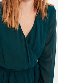 Robe enveloppante turquoise avec un décolleté en V et de longues manches transparentes. Un tissu doux qui tombe délicatement, avec une taille froncée et un éclat subtil.