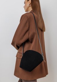 Bolso cruzado de ante negro con forma de media luna y herrajes plateados, combinado con un blazer marrón de gran tamaño.