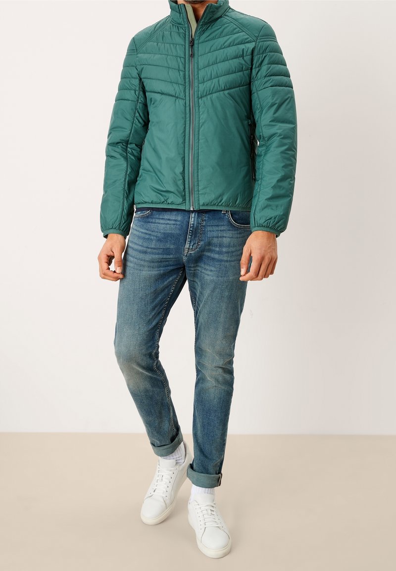 Homme portant une veste matelassée sarcelle, un jean bleu retroussé, des chaussettes blanches et des baskets blanches décontractées, debout sur un fond uni.