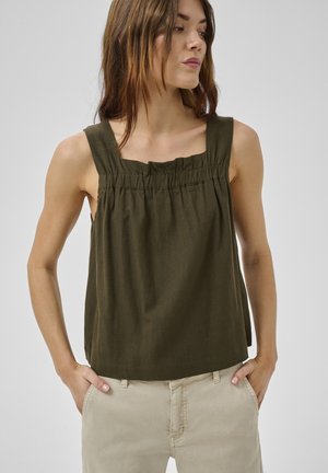 Žena s dlhými hnedými vlasmi, oblečená v olivovo zelenom tielku bez rukávov a béžových nohaviciach, stojaca so zavesenými rukami v vreckách, pozerajúca sa do strany.