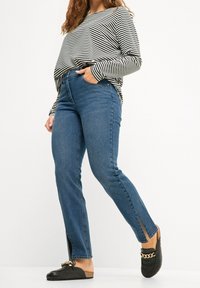 Top de manga larga a rayas en blanco y negro, jeans de tiro alto azules con aberturas laterales, y zapatos negros de tipo slip-on con detalle de cadena dorada.