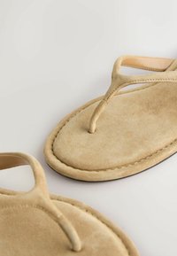 Sandales tongs en daim beige avec un design simple à brides et un léger rembourrage sur la semelle, présentées sur une surface gris clair.