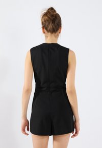  combinaison noire sans manches avec une taille cintrée et des shorts courts, dotée d'un col haut et d'une fermeture éclair au dos. Texture du tissu lisse.