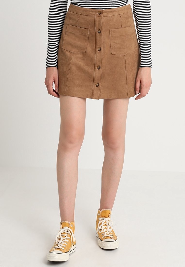 Bruine suède mini rok met twee voorkzakken, knoopsluiting en A-lijn ontwerp. Gecombineerd met gele hoge sneakers.