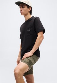 Νέος άνδρας που φοράει μπεζ καπέλο, μαύρο t-shirt Patagonia με τσέπη στο στήθος και χακί σορτς, ποζάροντας μπροστά σε απλό φωτεινό φόντο.