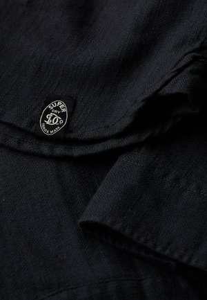 Tissu noir avec un motif en chevrons, comportant des accents brodés et une étiquette de marque circulaire noir et blanc.