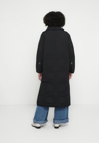 Black quilted long coat with a geometric pattern, high collar, and two side pockets, worn over light blue wide-leg jeans and white shoes.