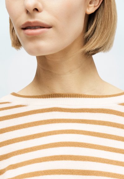 Rövid szőke hajú nő, bézs és fehér csíkos pulóverben, az állától a válláig látható, világos háttér előtt.