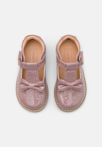 Różowe lakierowane buty z zapięciem T-strap, perforowanymi detalami i kokardką z przodu. Posiadają beżową podeszwę i wnętrze.