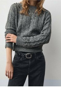 Grauer Strickpullover mit Kabelmuster, verziert mit metallischen Nieten an den Schultern und Bündchen, kombiniert mit schwarzen Jeans und einem Gürtel.