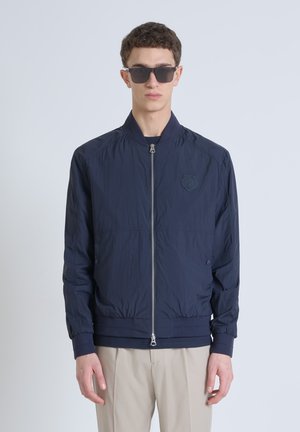 Giacca bombardiere blu navy con chiusura a zip e polsini a coste. Logo ricamato sul petto. Indossata sopra una maglietta scura e pantaloni chiari.