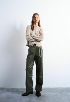 Ung kvinde står med armene krydsede iført beige strikket cardigan, mørkegrønne læderbukser med vide ben og mørkebrune sko foran en hvid væg.