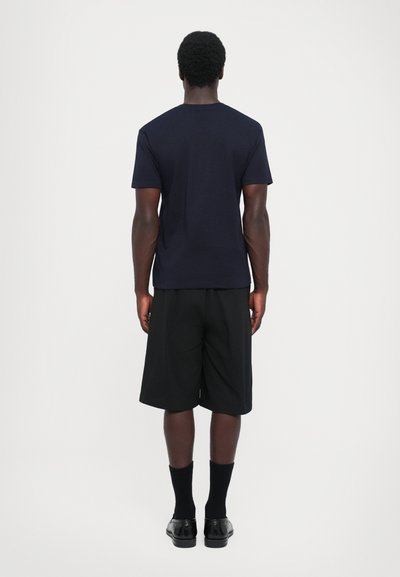 Navy bomulls T-skjorte med korte ermer, sammen med svarte knelange shorts. Person står med ryggen til, på en lys bakgrunn.