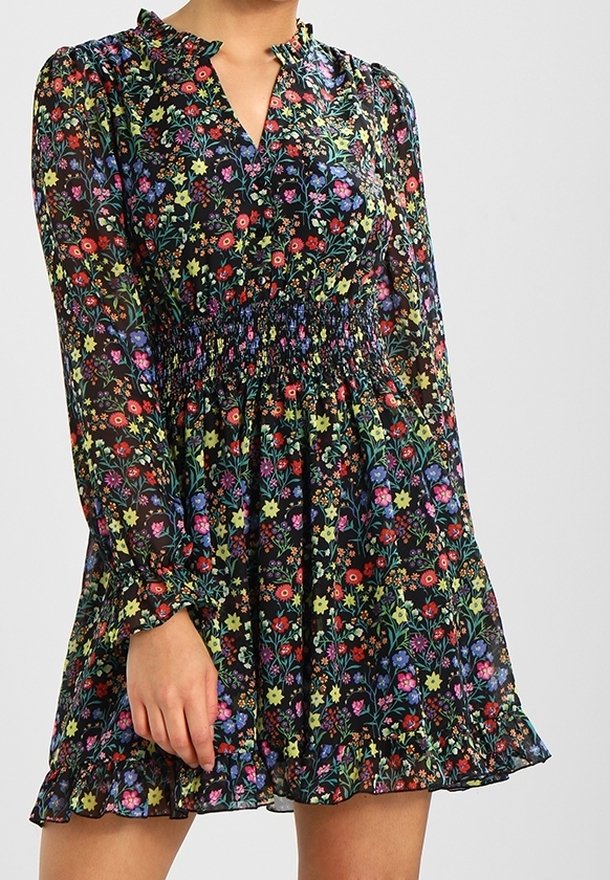 Femme portant une robe noire à motifs floraux avec manches longues, taille froncée, col à volants et ourlet à volants, debout devant un fond uni.