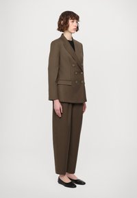 Bruine double-breasted blazer met grote knopen, gecombineerd met bijpassende losse broek en zwarte ballerina's, staande tegen een neutrale achtergrond.