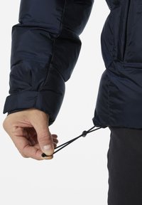 Chaqueta aislante azul marino con textura acolchada. Cuerdas ajustables en el dobladillo sostenidas por una mano, con topes redondos.