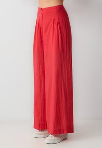 Pantalons larges rouges en tissu léger, dotés d'une taille haute, de plis sur le devant et d'une fermeture éclair à l'arrière. Assortis à des baskets blanches.