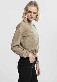 Beige bomullsgenser med camouflage-mønster, langermet og cropped, med høy hals og elastiske mansjetter, kombinert med svarte bukser.