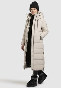 Lange beige pufferjas met een capuchon, verticaal gestikte patroon, zwarte ritsen, zijzakken, gecombineerd met zwarte laarzen en een gebreide muts.