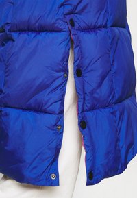 Blue puffer vest with a smooth, shiny texture. Features black snap buttons and contrasting pink lining at the opening. Sections are quilted.