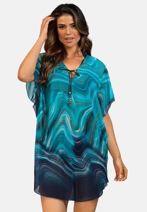 Kobieta ubrana w luźną, krótką sukienkę w turkusowo-granatowy wzór, z wiązaniem przy dekolcie, pozująca z jedną ręką przy włosach.