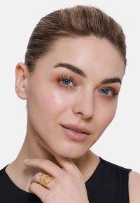Goldring mit verwobenen Strängen, der an einer Hand getragen wird. Das Modell hat helle Haut, blaue Augen und trägt ein schwarzes Oberteil vor einem weißen Hintergrund.