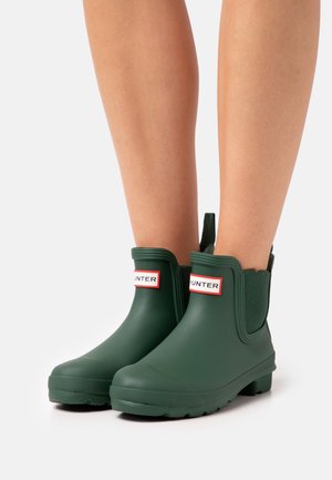 Bottes basses en caoutchouc vert à surface texturée, avec une étiquette de marque rouge et blanche. Le design comprend des panneaux latéraux élastiques et une semelle épaisse.