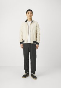 Giubbotto bomber leggero beige con dettagli neri, indossato sopra una t-shirt bianca. I pantaloni neri e le sneakers nere con lacci beige completano il look.