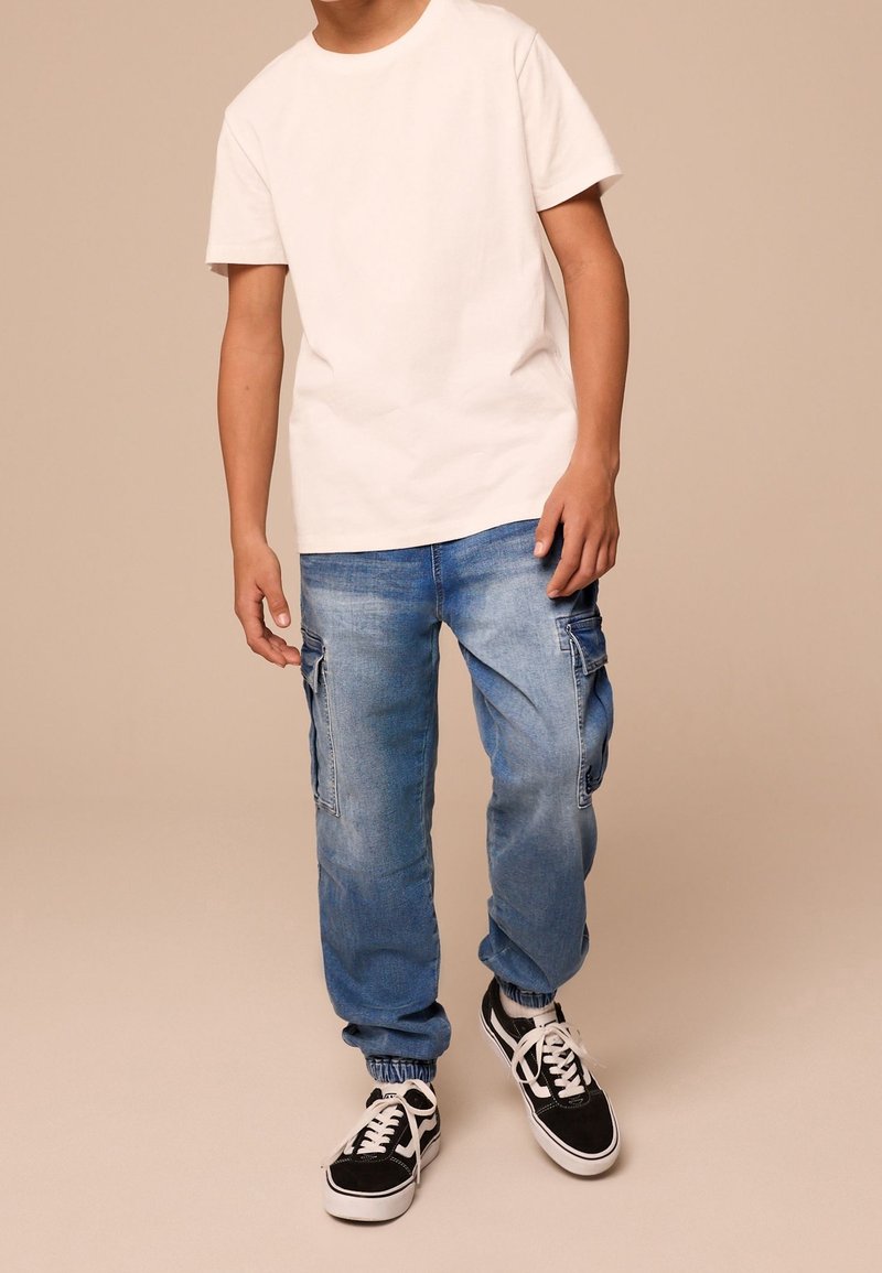 Jeune personne portant un t-shirt blanc uni, un jean cargo bleu clair avec des poignets élastiques et des baskets noires et blanches à lacets.