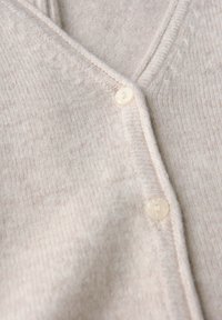 Närbild av beige stickad tyg med två krämfärgade runda knappar på kanten av en strukturerad cardigan.