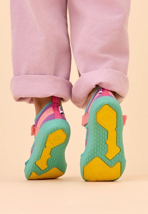 Person wearing pink rolled-up pants and colorful sandals with teal and yellow soles, standing on beige background.