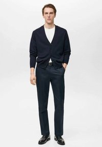 Mornarsko pleteno kardigan s petimi gumbi, bela majica s krožnim ovratnikom in temne hlače. Gladka tekstura, klasičen kroj in preprost dizajn. Črne čevlje.