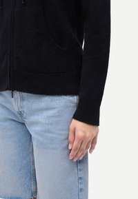 Svart zip-up-hoodie i mjukt material med framfickor, bärs över ljusblå denimjeans. Närbilden visar en hand vilande på benet.