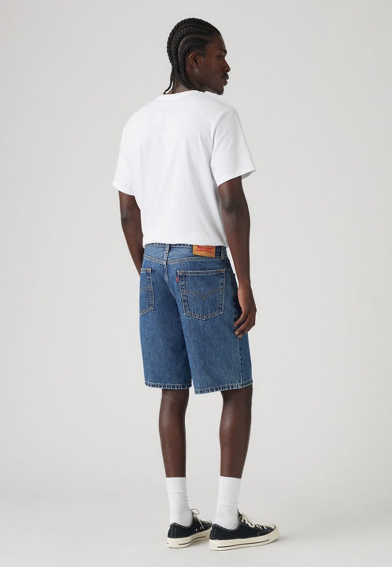 Homme aux cheveux tressés portant un t-shirt blanc, un short en jean bleu, des chaussettes blanches et des baskets noires, debout devant un fond uni.