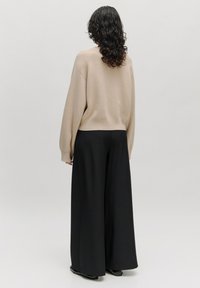 Beige gestrickter Pullover mit lockerer Passform, versehen mit überschnittenen Schultern, kombiniert mit schwarzen weiter geschnittenen Hosen aus einem glatten Stoff.
