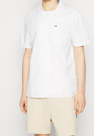Person trägt ein weißes kurzärmeliges Hemd mit kleinem Logo auf der Brusttasche und beige Shorts.