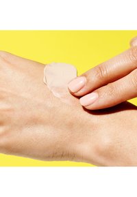 Beige cream on a hand, smooth texture, applied with fingers. Light pink nail polish on fingers. Bright yellow background.