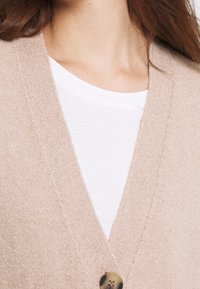 Béžový pletený kardigan s výstřihem do V, který má výrazné zapínání na knoflíky a měkkou texturu, vrstvený přes bílé tričko.