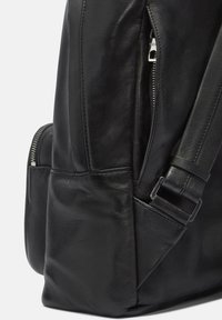 Czarny skórzany plecak o gładkiej fakturze, z zamykanymi na zamek kieszeniami i regulowanym paskiem. Charakteryzuje się ustrukturyzowanym kształtem i subtelnymi detalami przeszycia.