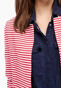 Rood-wit gestreepte cardigan van zacht materiaal, met een open voorkant. Gedragen over een marineblauw overhemd met knopen en zakdetails.